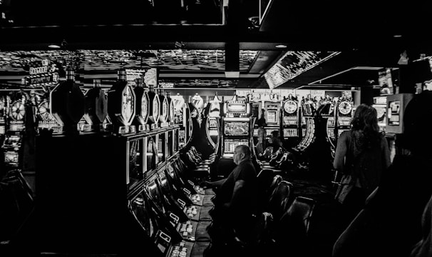 A dimly lit casino filled with rows of slot machines, some occupied by people. The machines' lights contrast with the dark surroundings, creating a vibrant yet subdued atmosphere.