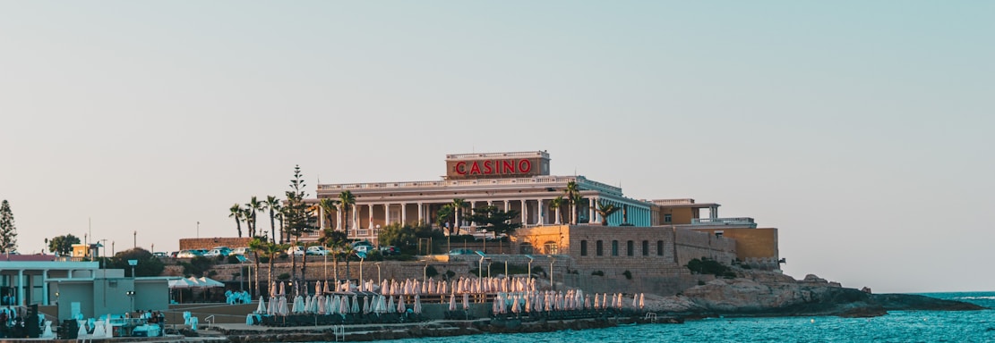 A seaside view of a large building labeled as a casino, situated on a rocky shore. The building is surrounded by palm trees and other smaller structures. The calm sea water reflects the sky, and there are several closed umbrellas lined along the waterfront.