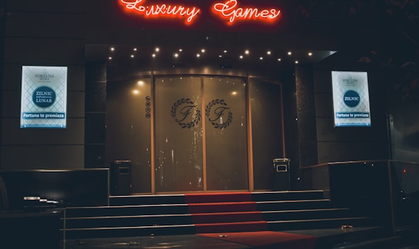 A neon-lit casino entrance with a red carpet leading up to large double doors. The sign reads 'Fortuna Palace Luxury Games' in bright red and white neon lights. The dimly lit atmosphere suggests a night-time setting. There are two digital displays on either side of the entrance showing promotional content.